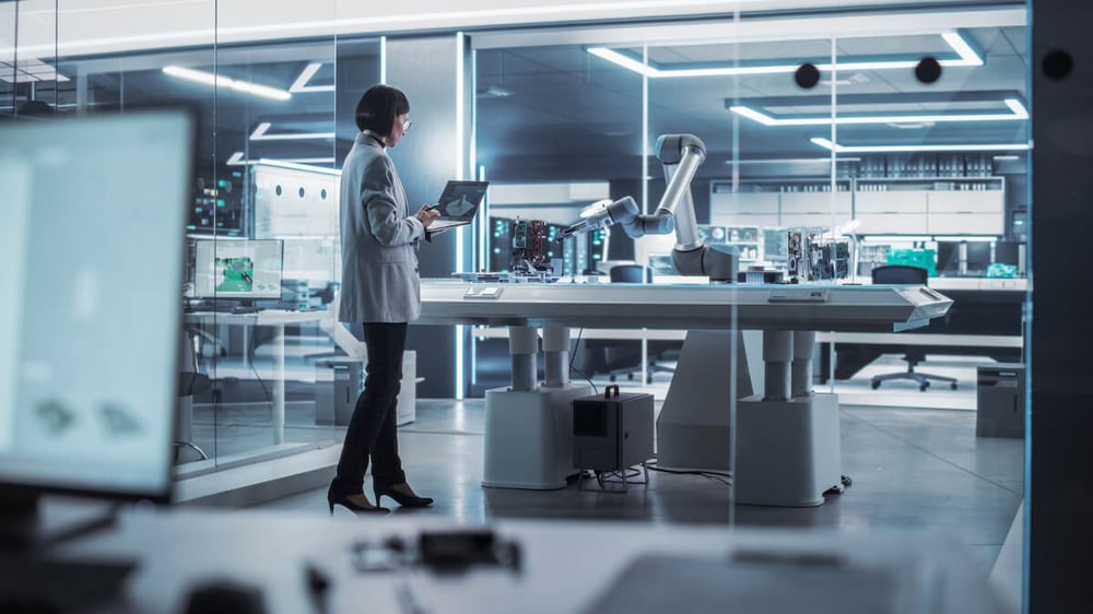 Female engineer standing next to a robotic arm holding a laptop with a prototype on screen.